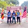 Three Rotary Cancer Run participants in event vests standing before the event backdrop, flanked by the Ugandan and Malaysian flags