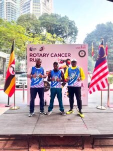 Three Rotary Cancer Run participants in event vests standing before the event backdrop, flanked by the Ugandan and Malaysian flags