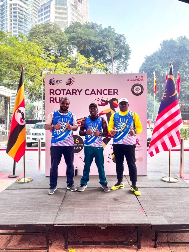 Three Rotary Cancer Run participants in event vests standing before the event backdrop, flanked by the Ugandan and Malaysian flags